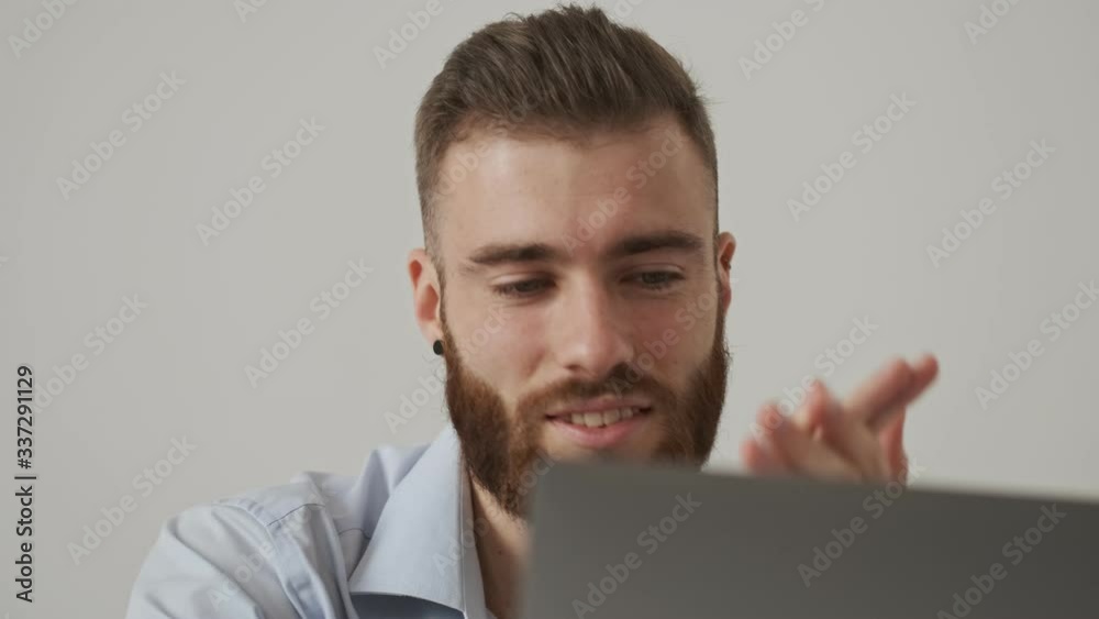 A satisfied glad young bearded man wearing a shirt is looking at his laptop computer at the white office A satisfied glad young bearded man wearing a shirt is looking at his laptop computer at the white office