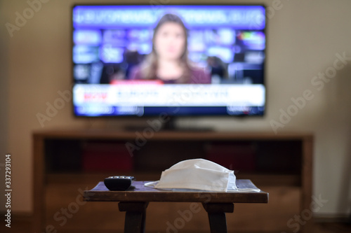 Stay at home concept in times of coronavirus pandemic, showing a television remote control and a medical mask, with news channel open in background