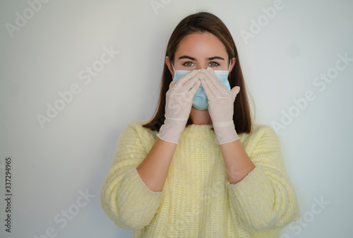 Happy smiling girl in sanitary gloves putting on medical face mask to protect herself from coronavirus during pandemic. White blank background and natural home light.