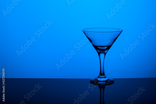 Glass goblet without wine on a thin leg stands on a mirror surface. Back light. Blue background. Copy space.