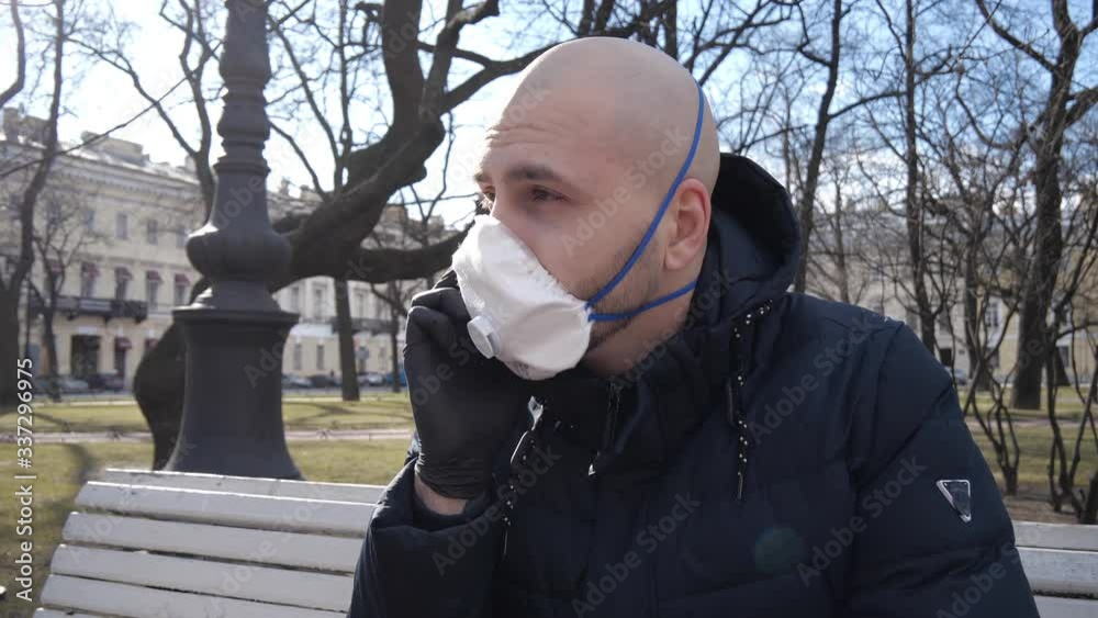 A young Caucasian guy with a bald head who has an infection is sitting in a respirator and medical gloves in a Park in the fresh air and discussing something on the phone A young Caucasian guy with a bald head who has an infection is sitting in a respirator and medical gloves in a Park in the fresh air and discussing something on the phone