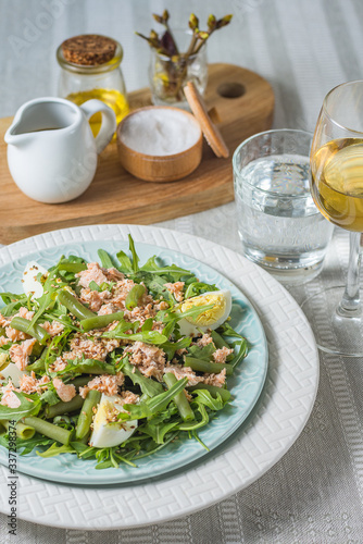 Arugula and trout salad with egg and asparagus on a plate on a table covered with a tablecloth and a glass of water and a glass of white wine vertical arrangement