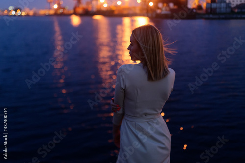 young lady in a beige dress hugging herself looks into the distance against the blue sea with lanterns