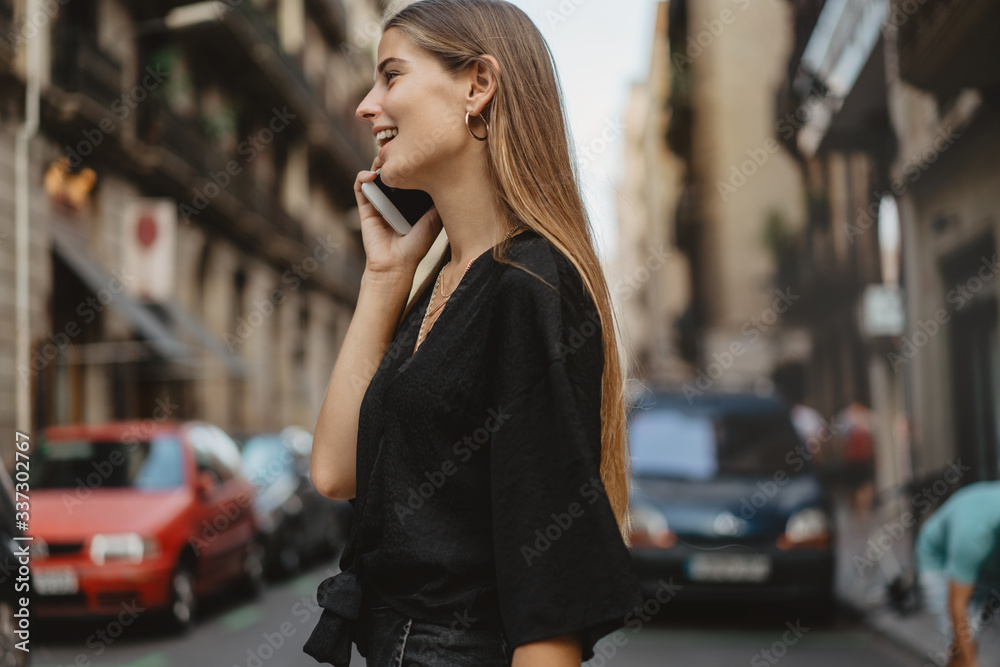 Fototapeta premium Girl accepts a call by mobile phone and starts an audio conversation in messenger with her friend while passing the road with cars walking in the city streets and enjoying the views