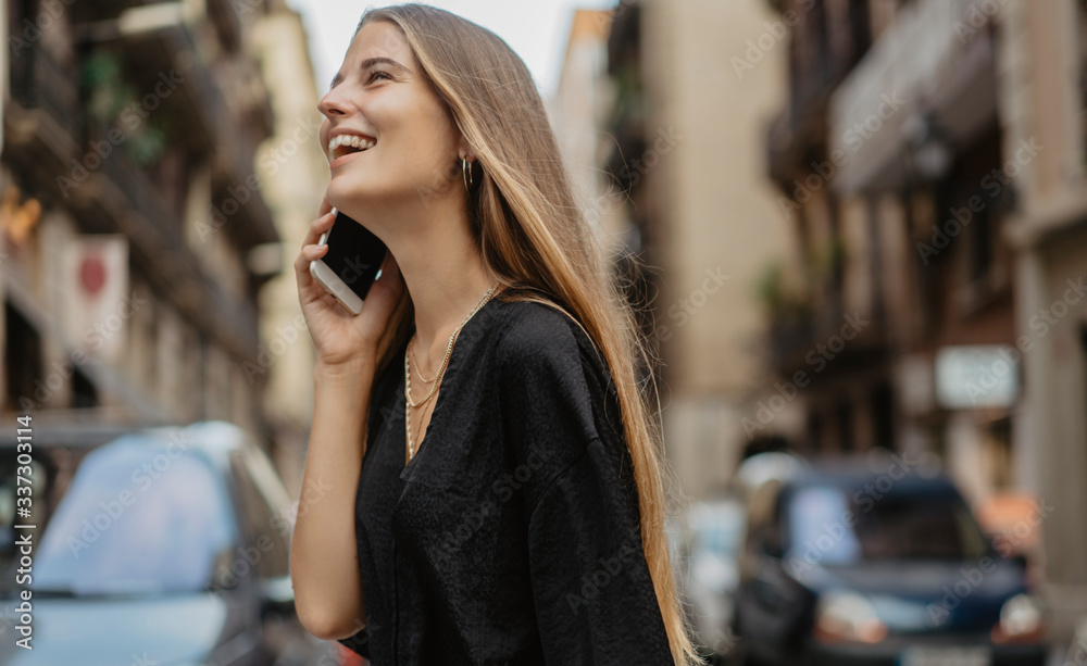 Young stylish girl with long blonde hair walking the streets of European city and making a phone call with her mobile cell phone, she is happy and laughing talking to her friend