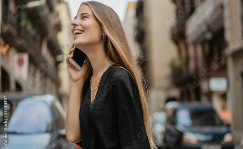 Young stylish girl with long blonde hair walking the streets of European city and making a phone call with her mobile cell phone, she is happy and laughing talking to her friend