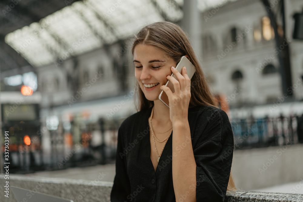 Young woman looks happy receiving some information by the call on her phone. She listen to the audio message while sitting at the railway station and waiting for vacations with her friends. Young woman looks happy receiving some information by the call on her phone. She listen to the audio message while sitting at the railway station and waiting for vacations with her friends.