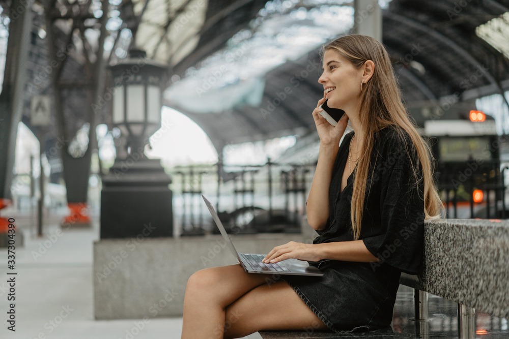 Girl is holding her smartphone making a call. Young blonde woman sitting on the stone bench and using her grey laptop for chatting with friends. She is waiting for the train at the railway station.