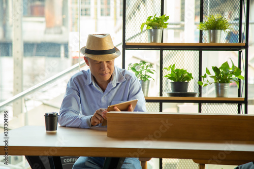 portrait middle aged Asian man in casual light blue shite and jeans pant and a cool hat sitting side coffee table bar using application on tablet with plant shelf with city urban building background.