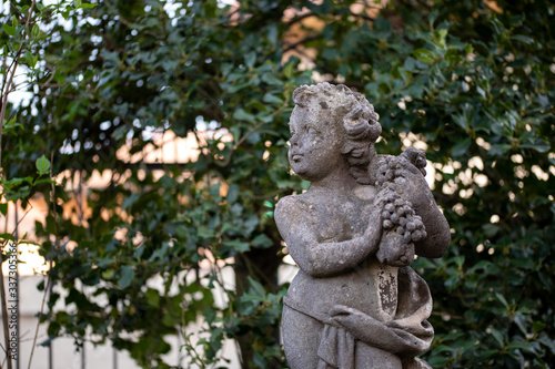 The courtyard statue decorations in a sunny day. The boy statue is standing look away. Close up white boy angel statue of white stone in cute gesture with blurred background of green plants in botanic