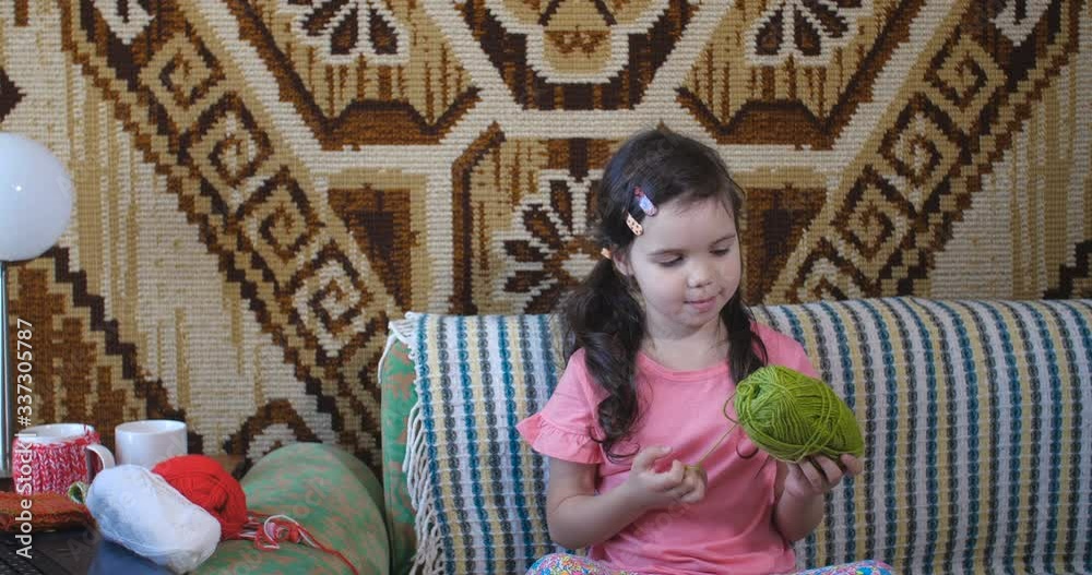 A little girl of five years old in pink T-shirt and with long brown hair sitting on sofa and playing with ball of green woolen threads. In background is woven Native American carpet with ornament. A little girl of five years old in pink T-shirt and with long brown hair sitting on sofa and playing with ball of green woolen threads. In background is woven Native American carpet with ornament.