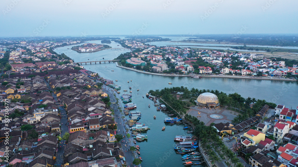 Aerial view of Hoi An old town on Thu Bon river at sunset, Quang Nam province, Vietnam. Unesco world heritage. Many boats moored to shore, bridges, Vietnamese houses on picture.