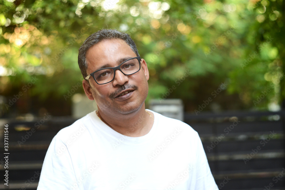 Indian man wearing spectacles in a white t-shirt outside a gate against a gate and a nature background Indian man wearing spectacles in a white t-shirt outside a gate against a gate and a nature background