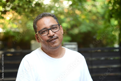 Indian man wearing spectacles in a white t-shirt outside a gate against a gate and a nature background