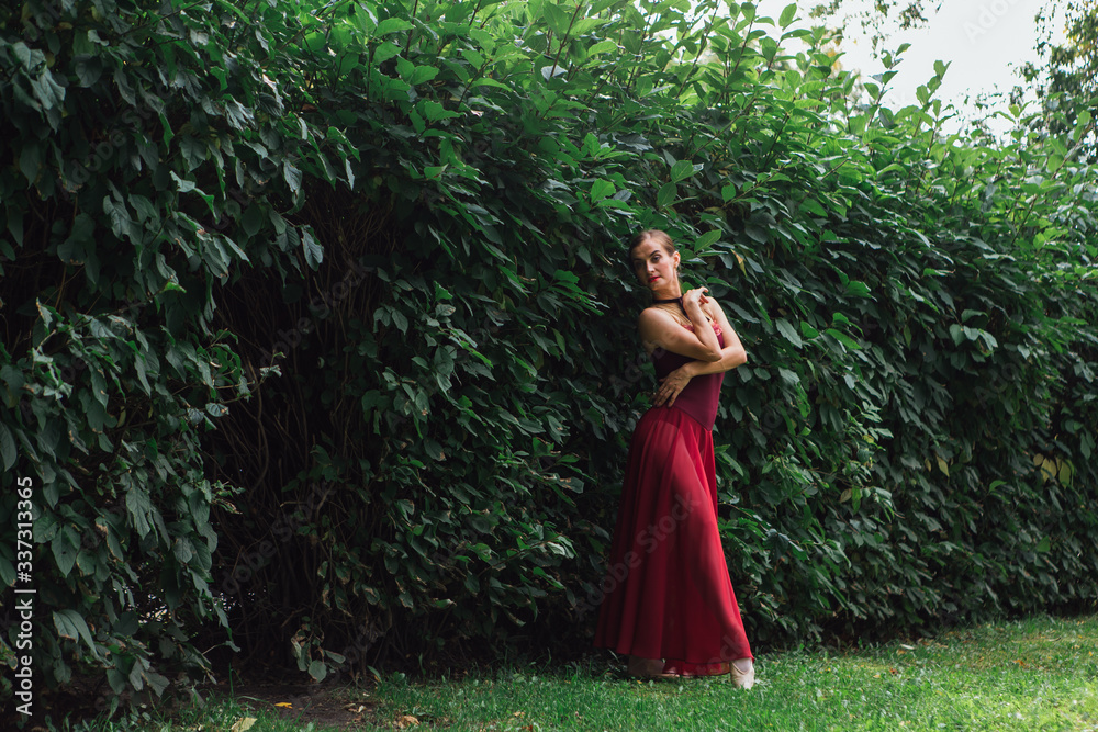 Woman ballerina in red ballet dress dancing in pointe shoes posing next to the wall of bushes Woman ballerina in red ballet dress dancing in pointe shoes posing next to the wall of bushes