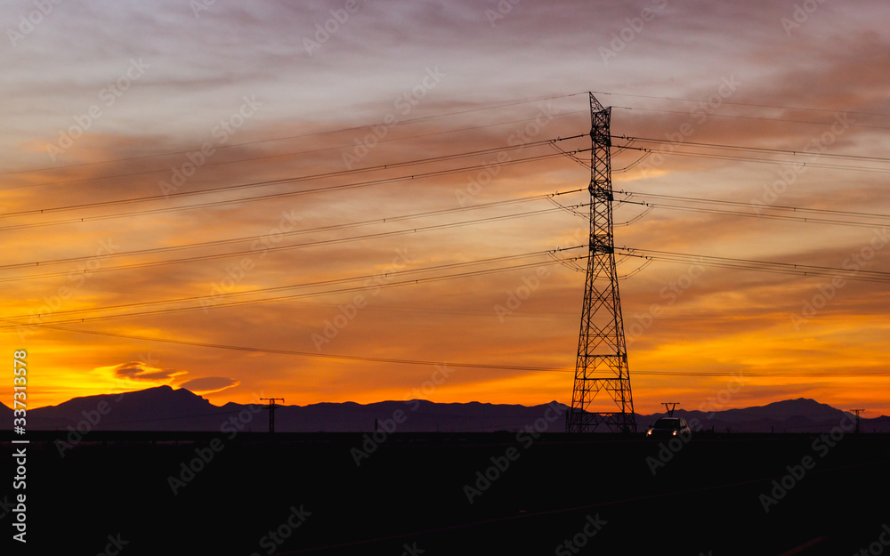 Silueta de tendido eléctrico con cables y torre de alta tensión en autovía sobre cielo anaranjado con nubes al sol del atardecer