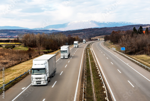 Convoy of White Trucks Or Traction Units In Motion On Road, Freeway. Asphalt Motorway Highway Against Background Of Mountains Landscape. Business Transportation And Trucking Industry.
