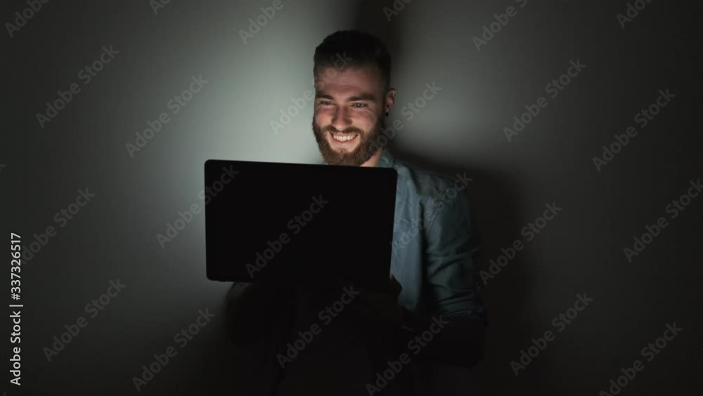 A handsome smiling young man wearing casual clothes is using his laptop computer at home leaning to the wall at night time A handsome smiling young man wearing casual clothes is using his laptop computer at home leaning to the wall at night time