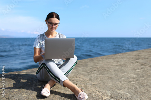 Beautiful young woman sitting with a laptop by the sea. Webinar online teaching concept. Online work.