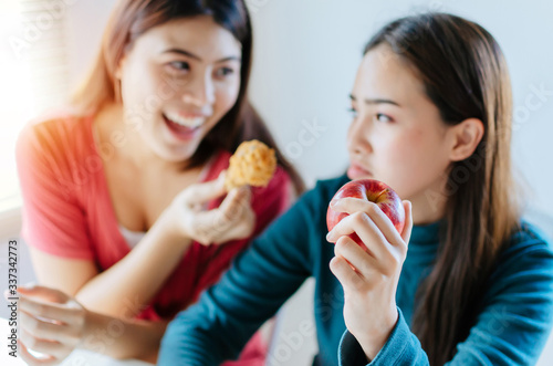 selective focus hand of young woman holding red apple and refuse to eat delicious hot fried chicken from friend smiling face at home  weight loss  diet plan  healthy lifestyle and dieting food concept