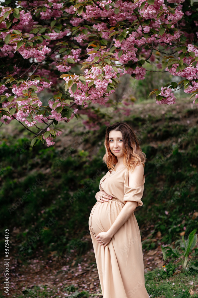 Young pregnant woman wearing beige dress standing near blossoming tree in spring. vertical portrait Young pregnant woman wearing beige dress standing near blossoming tree in spring. vertical portrait