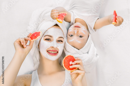 Happy family, mother and cute little daughter in white t-shirts and white towels on their heads lying on bed, doing facial beauty procedures with clay mask and holding slices of grapefruit in hands