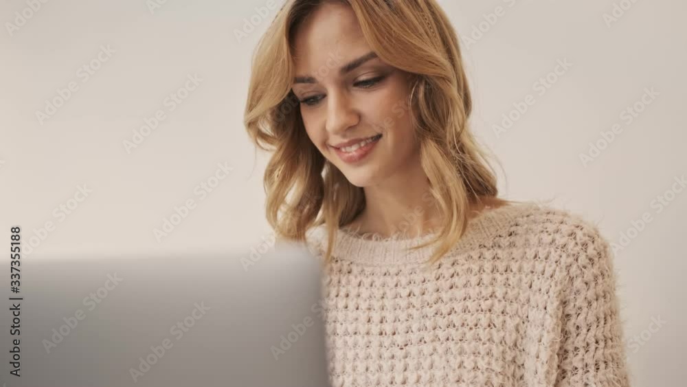 A young pleased woman is working with her silver laptop computer while sitting in the white room A young pleased woman is working with her silver laptop computer while sitting in the white room