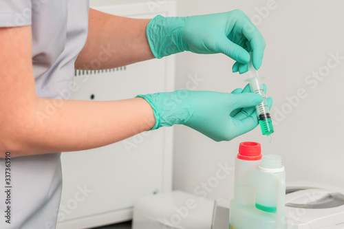 Hands in green gloves pours green liquid from syringe into plastic jar.