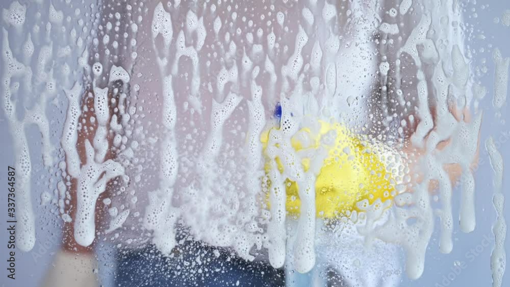 Woman wearing rubber glove spraying soap on window glass cleaning it with squeegee