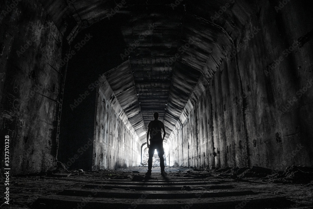 Shilouette of human underground explorer in abandoned dark tunnel with light in the end.