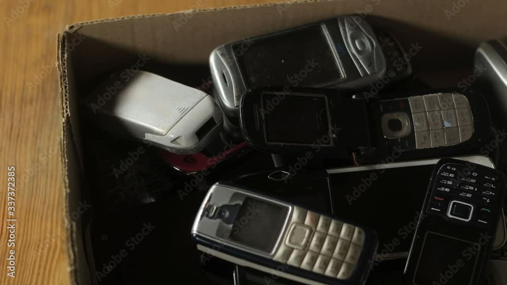 Outdated cell phones are in a big pile in a cardboard box. Old broken scratched mobile devices are recycled. analog communication. Outdated cell phones are in a big pile in a cardboard box. Old broken scratched mobile devices are recycled. analog communication.