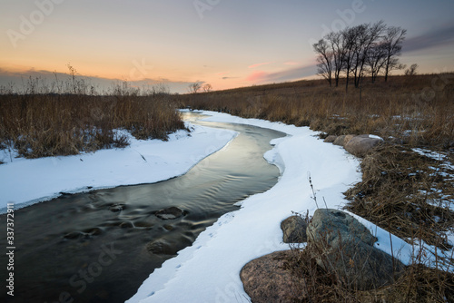 A scenic winter sunset over a stream flowing through a prairie landscape.
