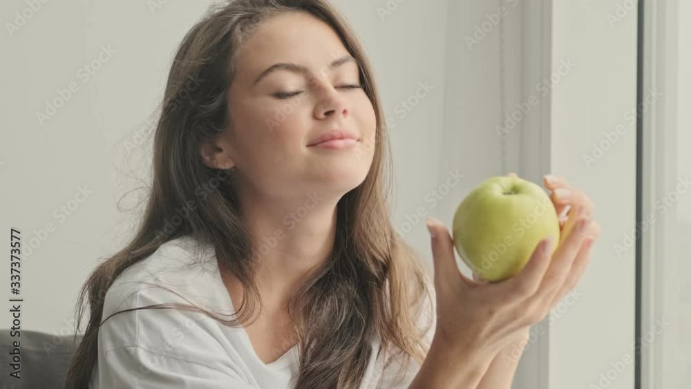 Pleased pretty brunette woman playing with apple and looking at camera while sitting on sofa at home Pleased pretty brunette woman playing with apple and looking at camera while sitting on sofa at home