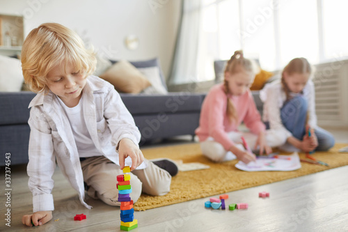 Little boy with blond hair playing on the floor with toys with his two sisters drawing in the background in the living room
