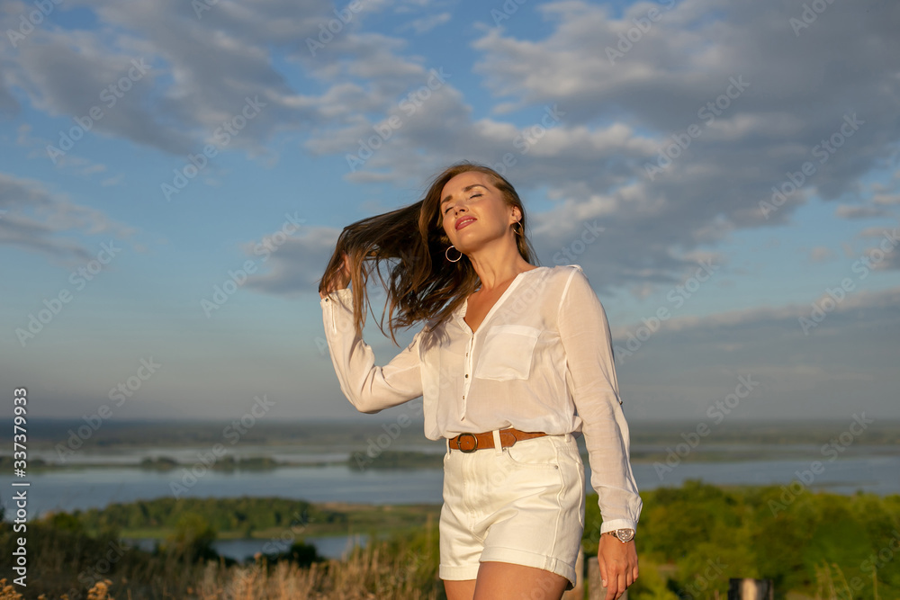 horizontal portrait of a long-haired European in a white blouse and white shorts against the backdrop of a beautiful river landscape horizontal portrait of a long-haired European in a white blouse and white shorts against the backdrop of a beautiful river landscape