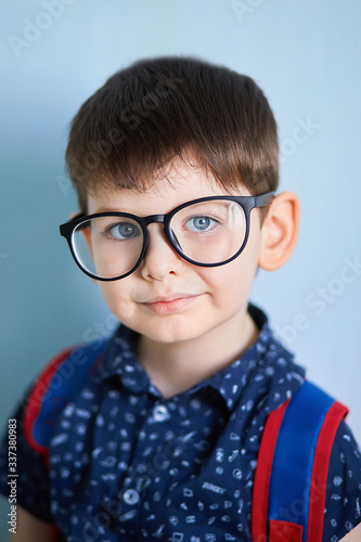 positive schoolboy with thumb up, in suit and glasses smiling and looking at camera while standing near table and browsing laptop against blue background