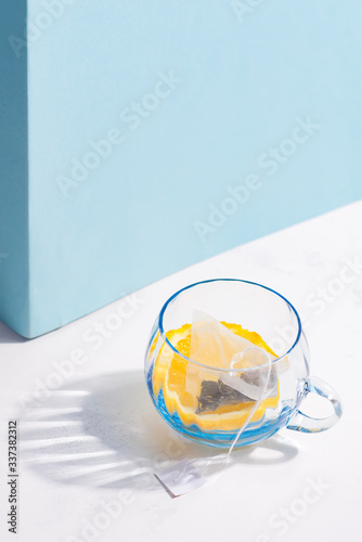 Glass cup with fruit tea bag and slice of lemon on a white table with shadow against pastel blue wall.