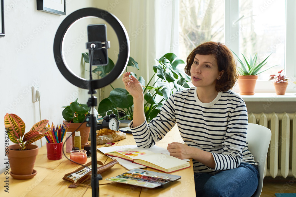 Female artist filming design workshop at home for online education. Working from home making master-class using ring lighting. Cozy artist's studio. Female artist filming design workshop at home for online education. Working from home making master-class using ring lighting. Cozy artist's studio.