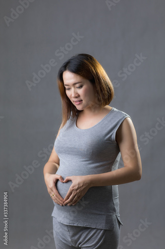 Pregnant woman in gray dress hold hands on belly in white background.