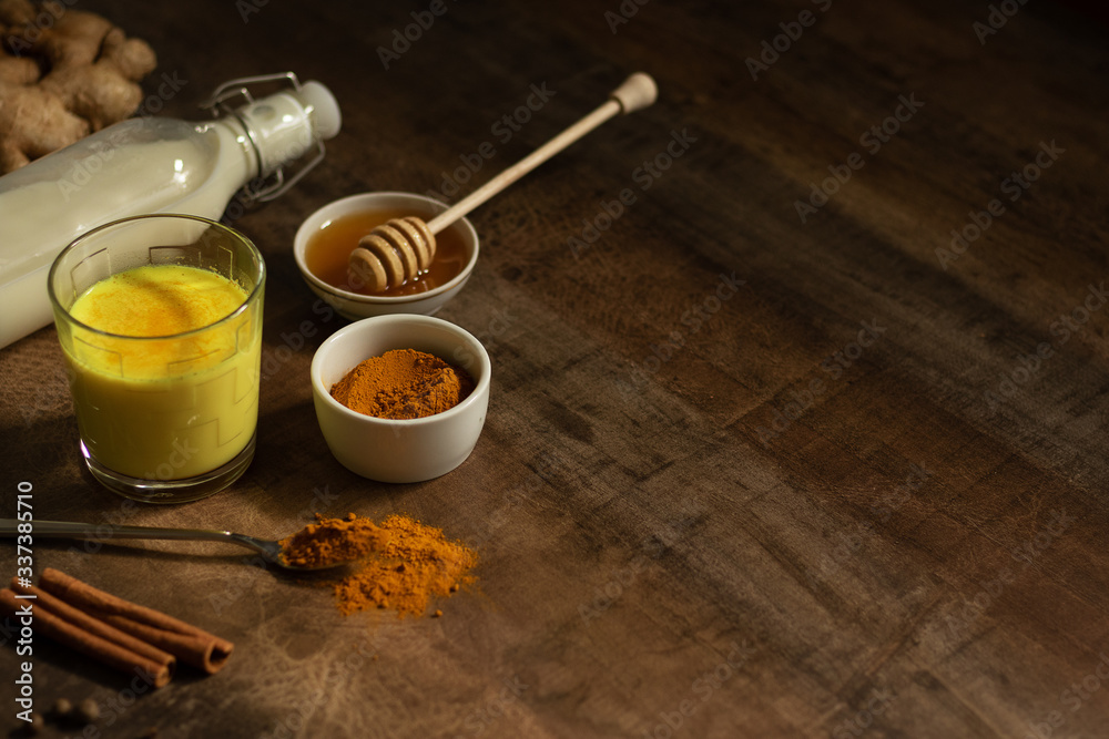Turmeric Milk is poured into a glass. Next to it are the ingredients for making a drink - milk in a bottle on a napkin, spices and honey. Golden drink on dark moody background, selective focus. Top Turmeric Milk is poured into a glass. Next to it are the ingredients for making a drink - milk in a bottle on a napkin, spices and honey. Golden drink on dark moody background, selective focus. Top