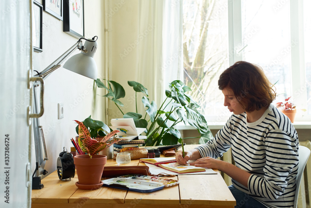 Female artist at her workplace working from home. Woman dressed in jeans and striped shirt sitting at the table and drawing illustration.