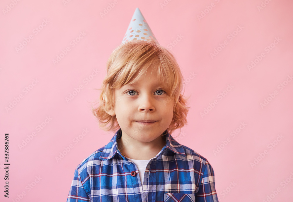 Portrait of little boy with blond hair and in party hat looking at camera over pink background Portrait of little boy with blond hair and in party hat looking at camera over pink background