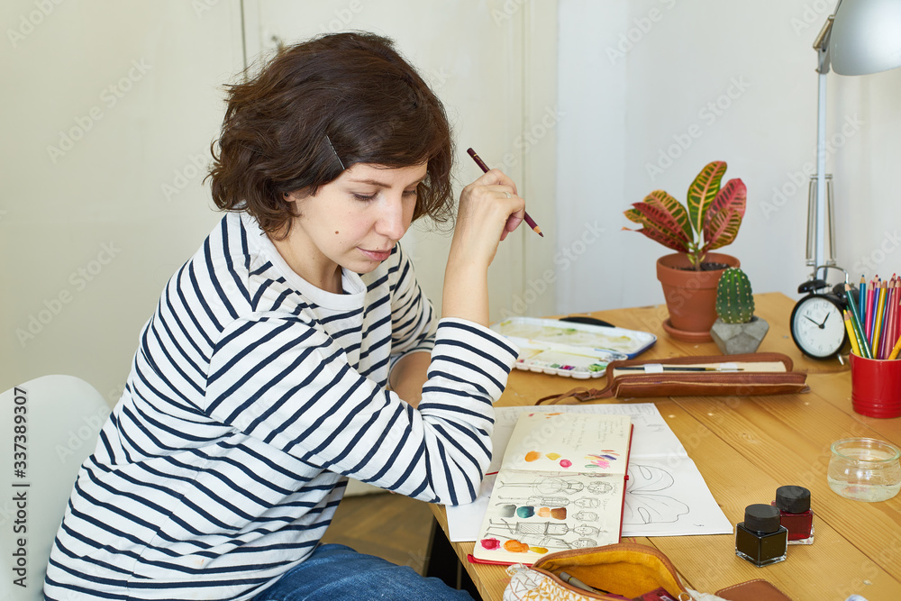 Female artist at her workplace working from home. Woman dressed in jeans and striped shirt sitting at the table and drawing illustration. Female artist at her workplace working from home. Woman dressed in jeans and striped shirt sitting at the table and drawing illustration.