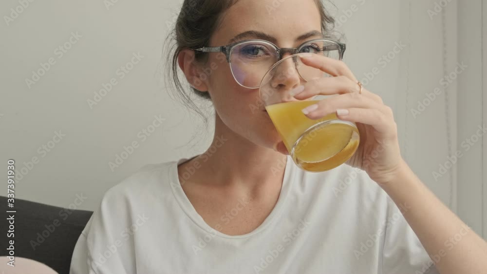 Happy pretty brunette woman in eyeglasses drinking juice and looking at camera while sitting on sofa at home