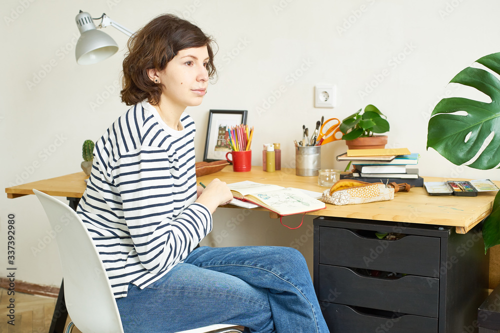 Female artist at her workplace working from home. Woman dressed in jeans and striped shirt sitting at the table and drawing illustration. Female artist at her workplace working from home. Woman dressed in jeans and striped shirt sitting at the table and drawing illustration.