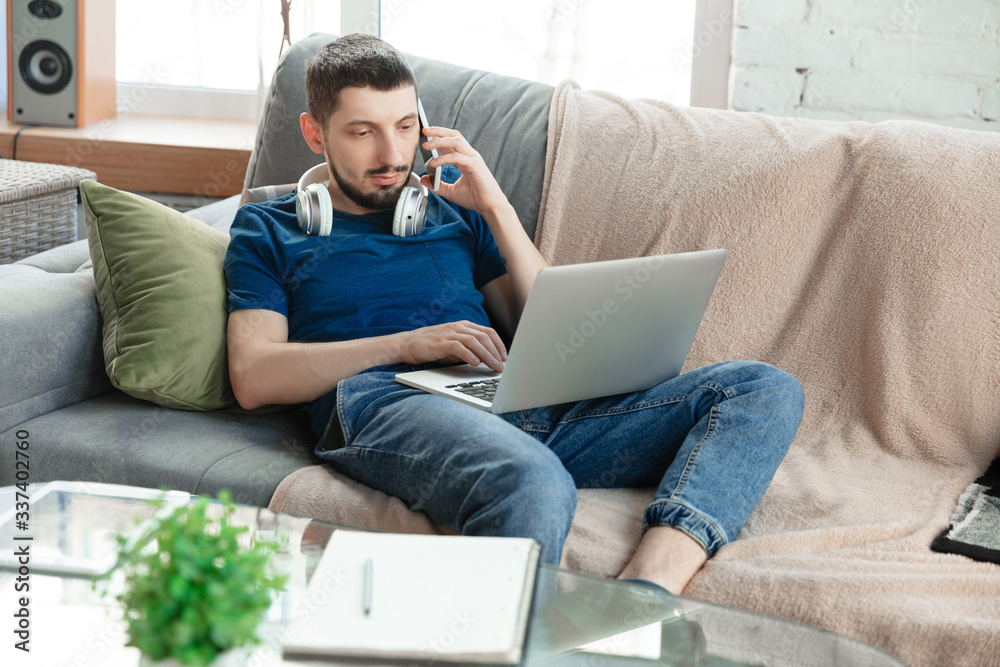 Young focused man studying at home during online courses or free information by hisself. Becomes software developer, IT manager while isolated, quarantined. Using laptop, smartphone, headphones. Young focused man studying at home during online courses or free information by hisself. Becomes software developer, IT manager while isolated, quarantined. Using laptop, smartphone, headphones.