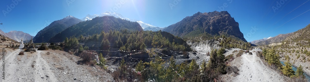 A panoramic view on Himalayas along Annapurna Circuit Trek, Nepal. There is a vast, dried plain around. Few prayer flags waving on the wind, river in the bottom