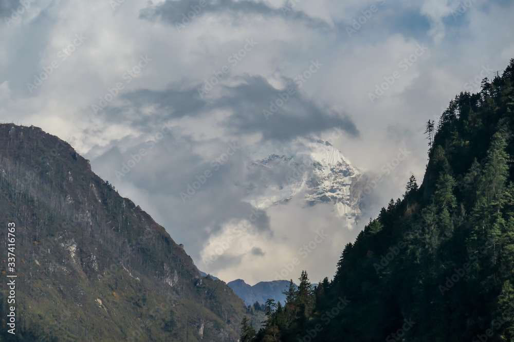 View on Manaslu hiding in the clouds from Annapurna Circuit Trek, Nepal. There is a dense forest in front. High, snow caped mountains' peaks catching the sunbeams. Serenity and calmness. Barren slopes