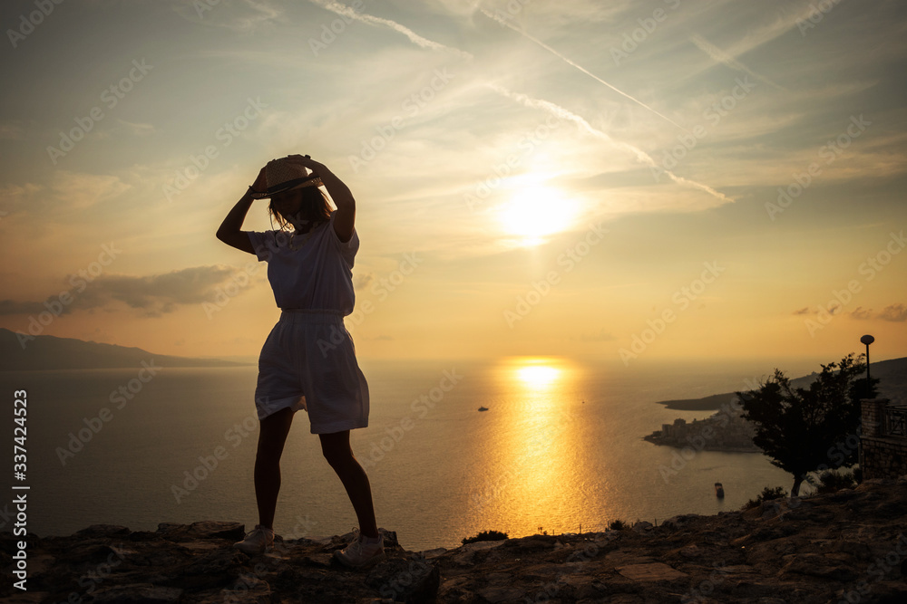 Fototapeta premium Young woman in hat, white t-shirt and shorts standing on the stone, looking at sunset over the sea.