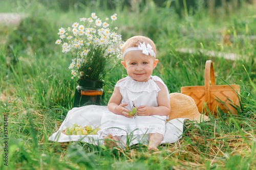 a little girl 1 year old sits in the grass in light clothes, holds grapes in her hand, smiles, next to her is a basket and a bouquet of field daisies, childhood in the summer